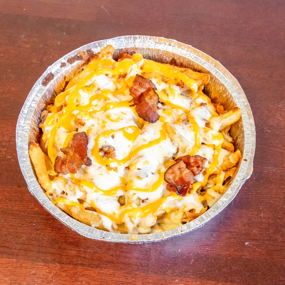 Loaded fries topped with melted cheddar, white cheese sauce, and crispy bacon in a foil tray on a wooden table.