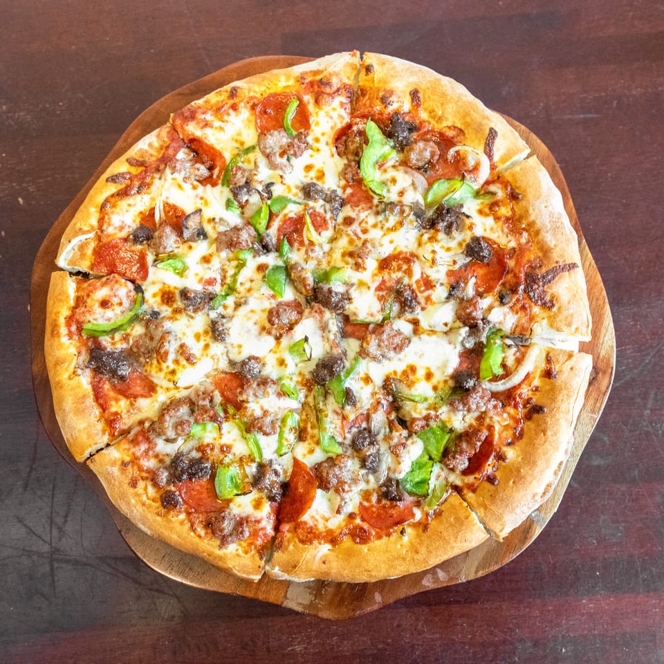 Large sliced pizza topped with melted cheese, pepperoni, sausage, mushrooms, green peppers, and onions on a wooden serving board.