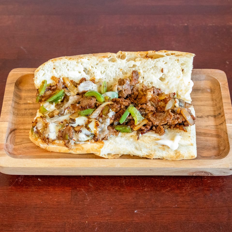Cheesesteak sandwich with sliced beef, melted cheese, peppers, onions, and mushrooms on a wooden serving board.