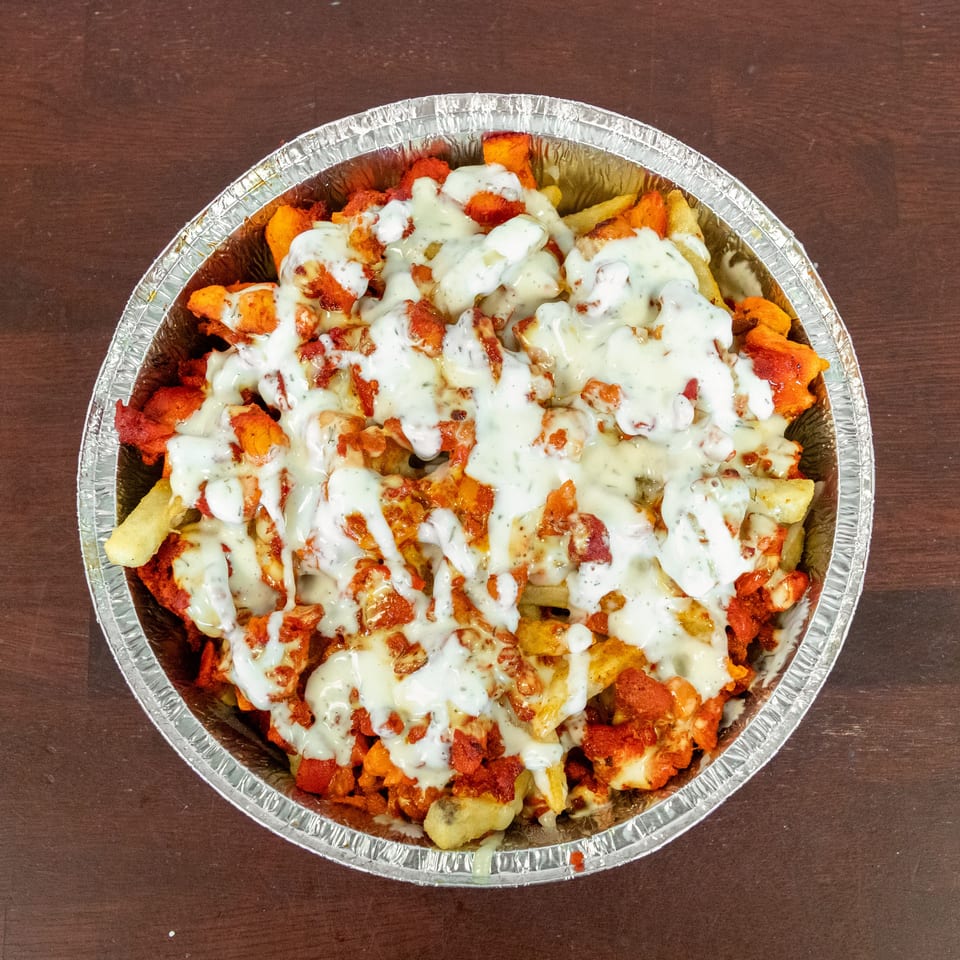 Loaded fries topped with seasoned chicken, melted cheese, and creamy white sauce in a foil tray on a wooden table.