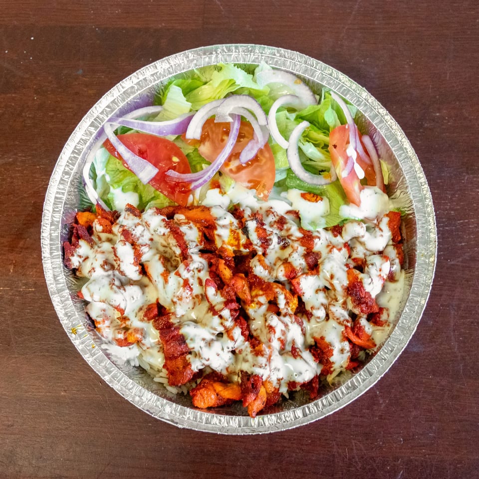 Aluminum bowl of seasoned chicken over rice, topped with creamy white sauce and served with lettuce, tomatoes, and onions.